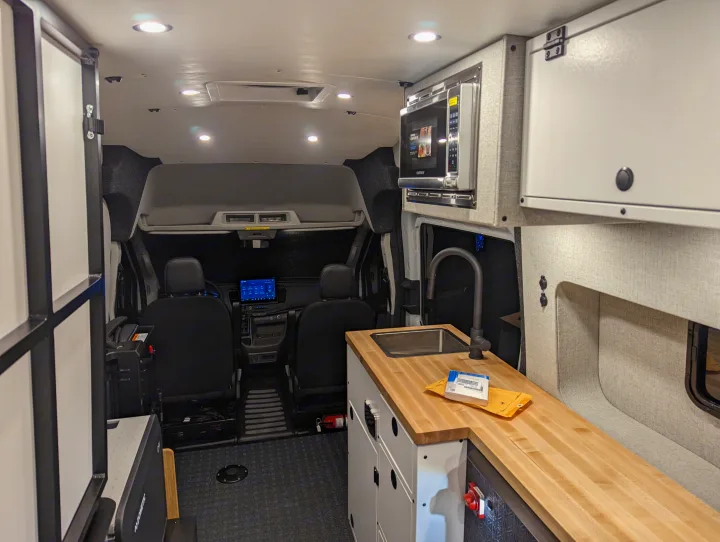 Inside an RV rental: a compact kitchen area with wood countertop, sink, microwave, and driver's cabin visible forward.. Grit Overland GO A/T 2022