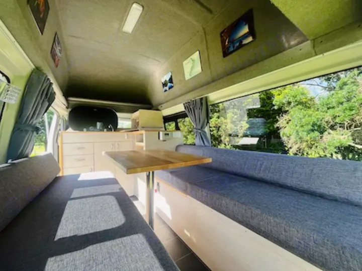 RV rental interior: large window offers tree views from comfy grey benches & dining table; compact kitchen visible.. Toyota Hiace 2014