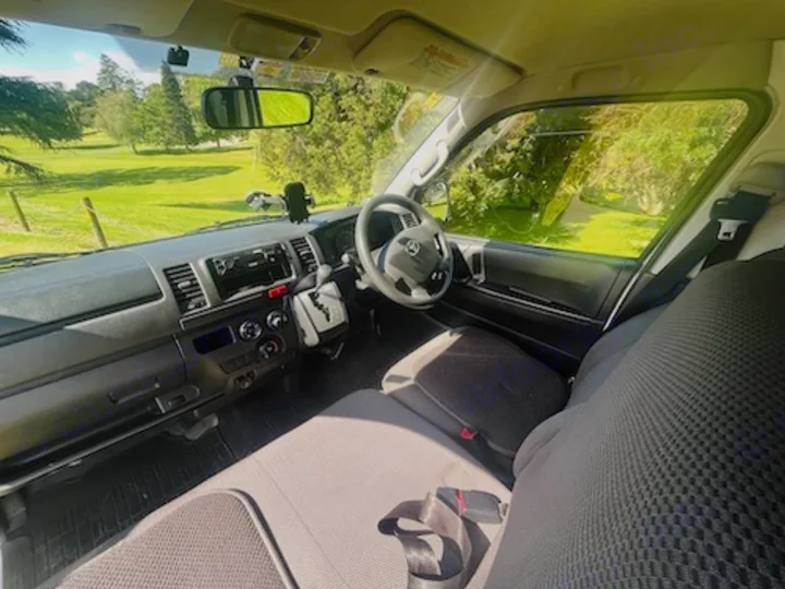 Inside an RV rental: a bright, sunny view from the driver's seat of the dashboard, seats, and green outdoors.. Toyota Hiace 2014