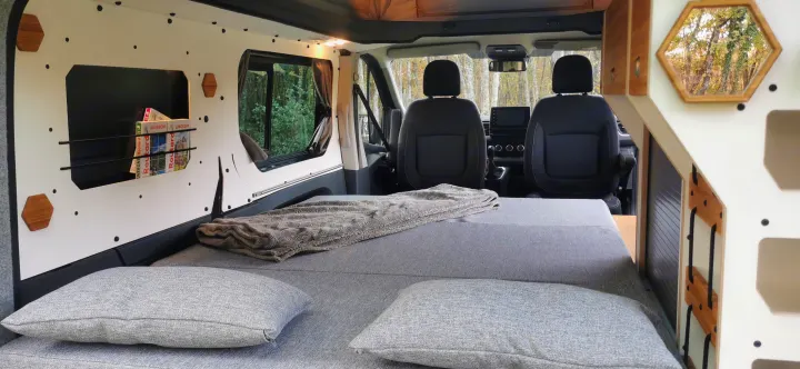 Step into this inviting RV rental interior, featuring a large gray bed with pillows, magazine storage, and front seats.. Renault Trafic 2023