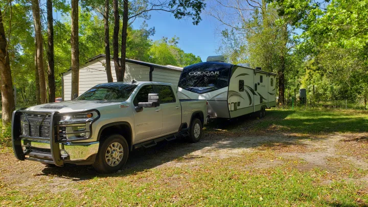 Silver truck towing an RV rental, parked on a dirt path under tall trees next to a metal shed on a sunny day.. Keystone RV Cougar 2020
