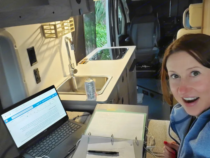 Smiling woman works remotely in her RV rental. Features a compact kitchen & mobile office for comfortable van life.. Winnebago Solis 2021