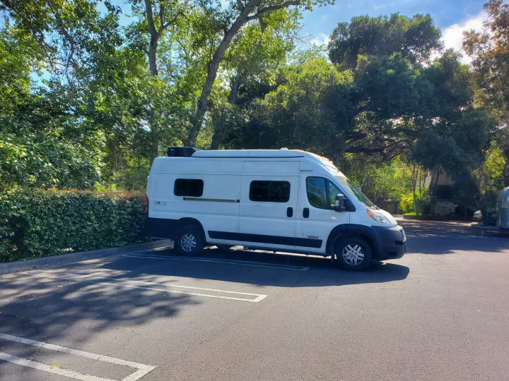 A white camper van, suitable for an RV rental, parked in a sunny lot with green trees.. Winnebago Solis 2021