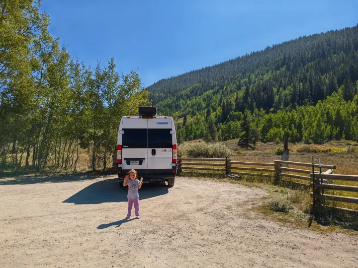 A child stands near a white RV rental (Winnebago Solis) parked on a dirt road in a sunny mountain forest.. Winnebago Solis 2021