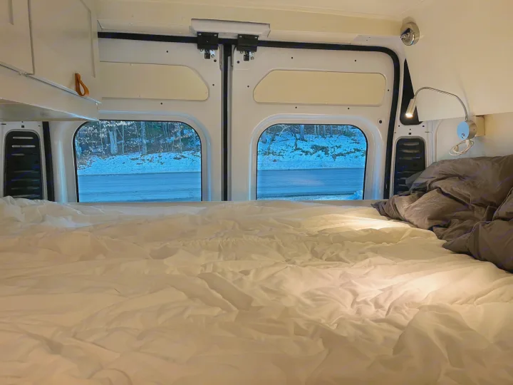 Cozy rv rental interior: A bed with white sheets and gray bedding, offering a view of a snowy road and forest for winter travel.. Ram Promaster 2022