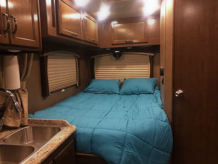 RV rental interior featuring a cozy bedroom with a blue comforter, two pillows, overhead cabinets, and a kitchen sink.. Thor Chateau 2017
