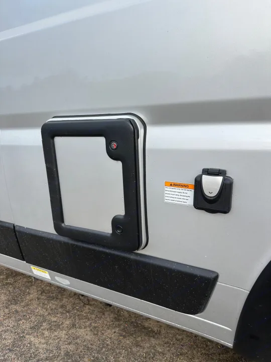 Side of an RV rental: external 30-amp electrical hookup with warning label and an access panel are visible on the silver exterior.. Winnebago Solis 2022