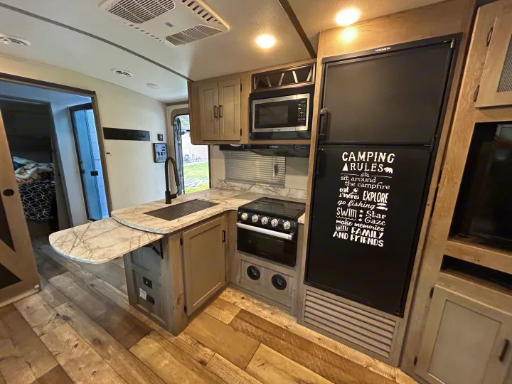 Spacious RV rental kitchen, featuring a sink, stove, microwave, & fridge with "Camping Rules" decal. Ready for travel adventures!. Keystone RV Outback Ultra-Lite 2021
