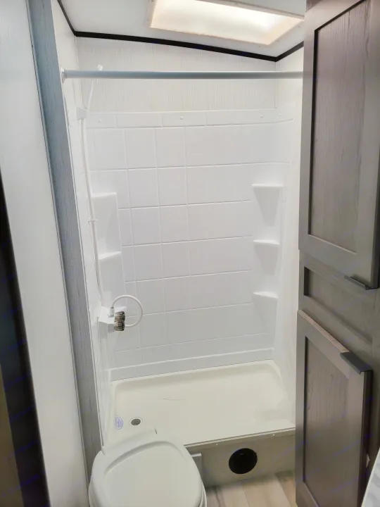 Inside an RV rental: Compact wet bath with white shower walls, a toilet, flexible shower head, and dark wood cabinetry.. Cruiser RV MPG Ultra Lite 2022