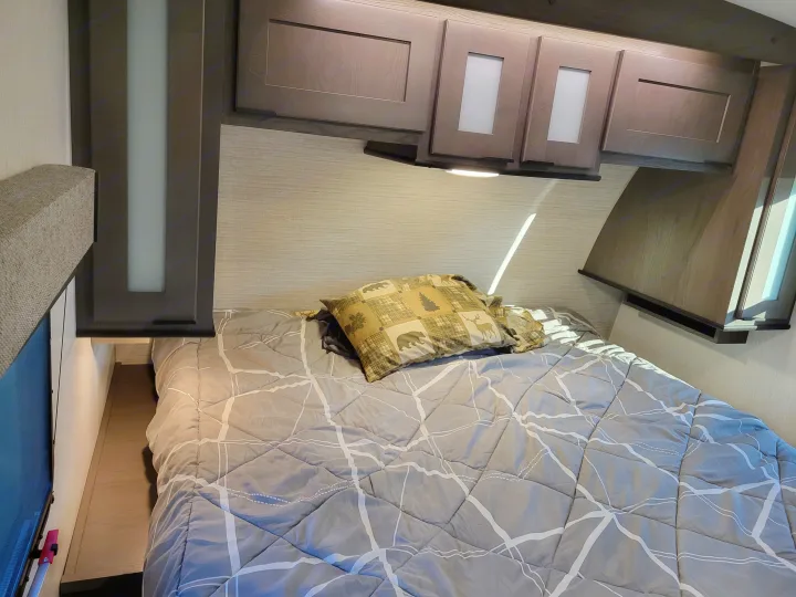 Inside an RV rental: a neat bedroom with a patterned gray comforter, rustic-themed pillow, and overhead wood cabinets.. Cruiser RV MPG Ultra Lite 2022