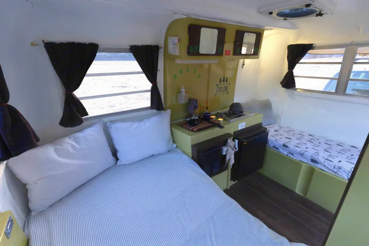 Cozy RV rental interior showcases a large bed, compact kitchen with stove & fridge, and a patterned single bunk.. Trillium Rv 1300 1976