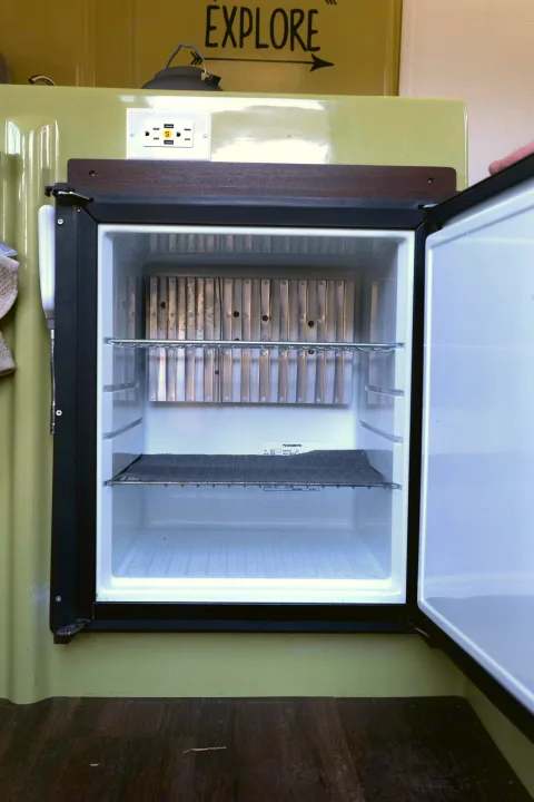 Empty and open compact refrigerator inside an rv rental, showing shelves, cooling fins, and an outlet above.. Trillium Rv 1300 1976