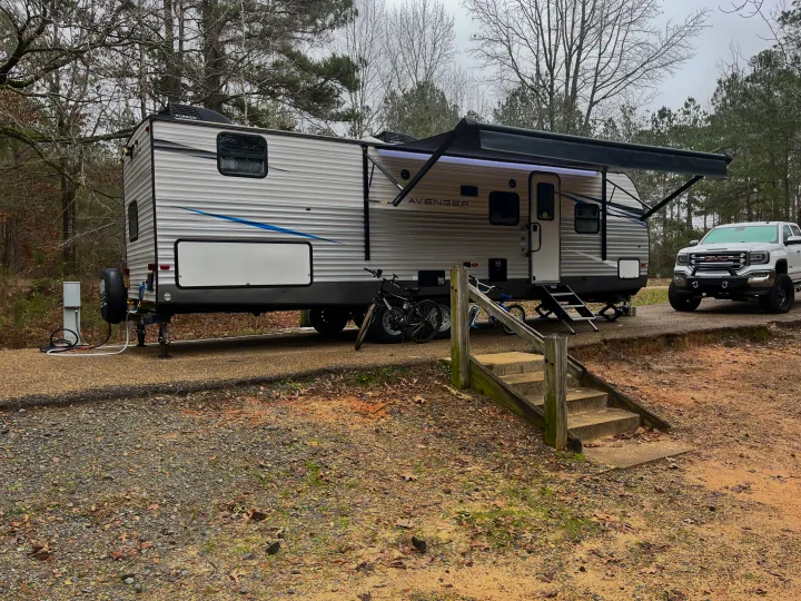 An RV rental, a large travel trailer, is set up at a wooded campsite with a white GMC truck and bikes.. Avenger Avenger Trailer 2021