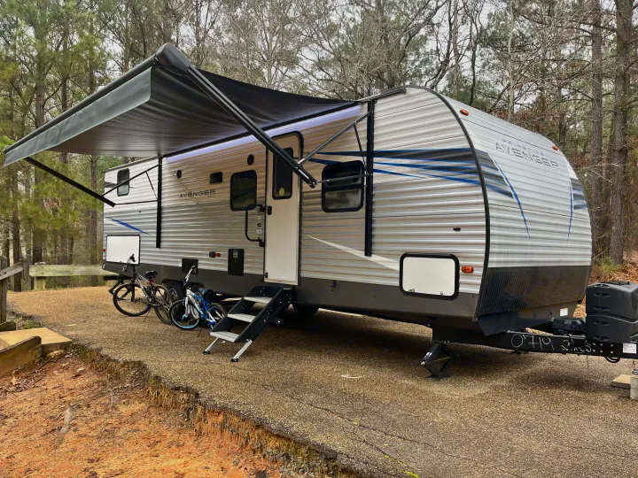This travel trailer RV rental, with its awning extended and bikes parked, is set up for outdoor adventure at a wooded campsite.. Avenger Avenger Trailer 2021