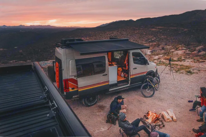 Friends by a campfire, bikes, and an rv rental camper van at sunset. Perfect for mountain adventures and outdoor camping.. Storyteller Overland Beast Mode 2023