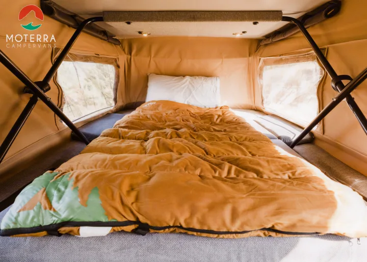 Interior of an RV rental: a cozy bed with a brown sleeping bag (green mountain design) & white pillow, surrounded by bright windows.. Mercedes-Benz Sprinter 2020