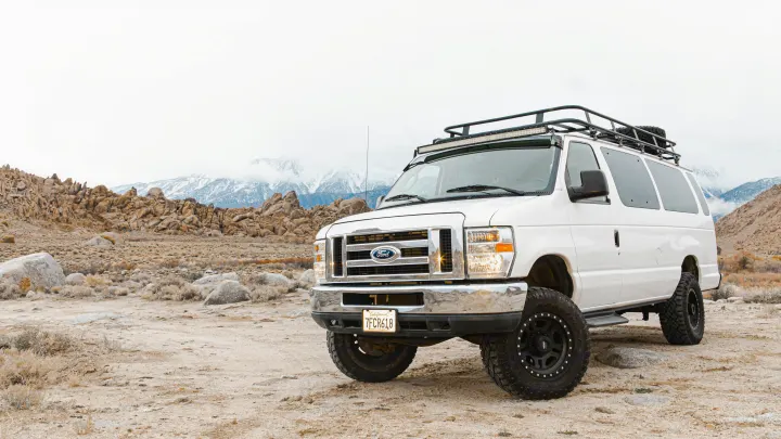 Rugged white Ford adventure van with roof rack & light bar in rocky desert. Snow-capped mountains. The perfect off-road RV rental.. Ford Ford E350 2014