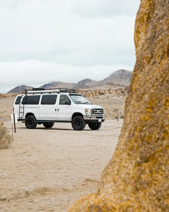 White off-road RV rental van with roof rack and ladder, ready for adventure in a rocky desert landscape.. Ford Ford E350 2014