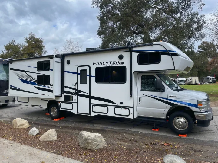 A white Class C RV rental is parked at a campsite with its slide-out extended, stabilized by orange leveling blocks.. Forest River Forester 3251DSF 2023