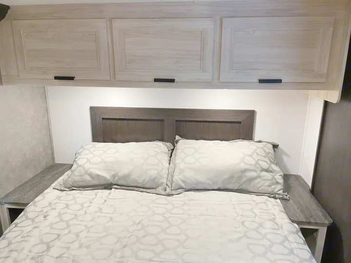 Cozy RV rental bedroom featuring modern grey bedding, a dark headboard, and light wood overhead cabinets for storage.. Forest River Forester 3251DSF 2023