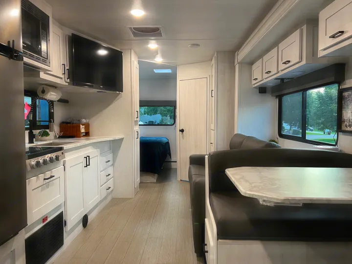 Bright RV rental interior with light wood floors, white cabinetry, a modern kitchen, dining booth, and a peek into the bedroom.. East to West Entrada 2600DS 2022