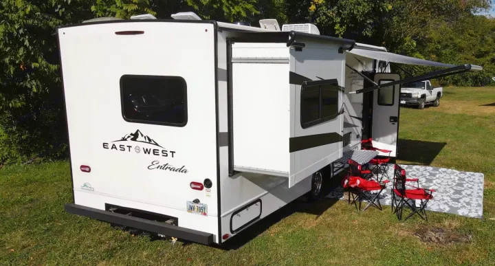 This white RV rental shows an extended slide-out and awning. Red chairs & a table sit on a patterned rug for outdoor camping.. East to West Entrada 2600DS 2022