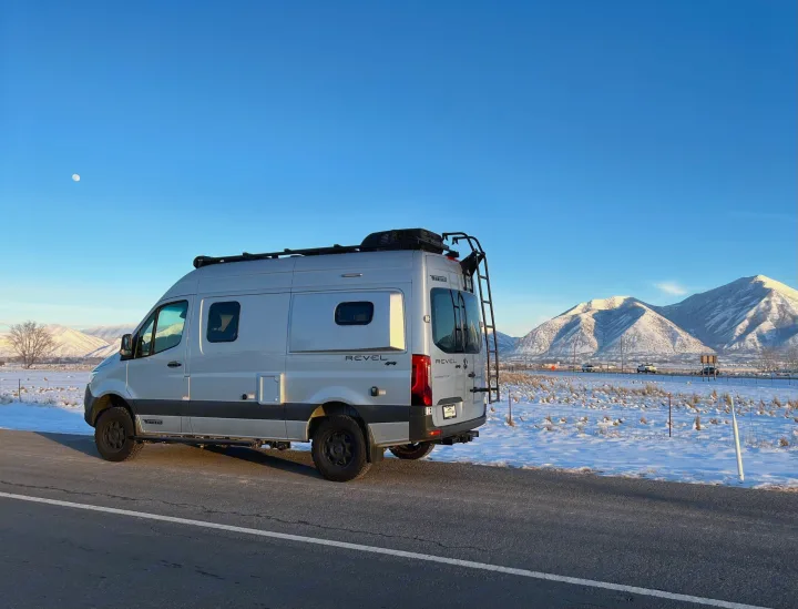 Silver Winnebago Revel camper van, ideal for rv rental, parked on a snowy roadside with stunning snow-capped mountains and a moon overhead.. Winnebago Revel 2020