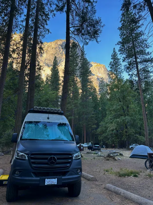 An RV rental Mercedes Sprinter campervan at a scenic mountain campground, surrounded by tall pine trees and a massive rock face.. Other Revel 2023
