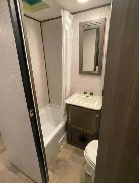 View of an RV rental bathroom with a white tub, shower curtain, sink vanity, mirror, and toilet.. Dutchmen Kodiak SE 2021