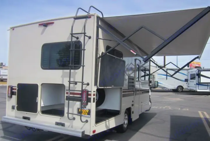 Cream-colored RV with an extended awning, open storage, and rear ladder, perfect for rv rental adventures.. Other Leprechaun 2022