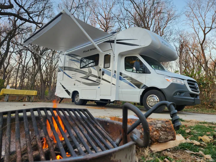 An RV rental campsite scene: a Thor Four Winds motorhome with awning extended, a picnic table, and a warm campfire in the foreground.. Thor Motor Coach Four Winds 2023