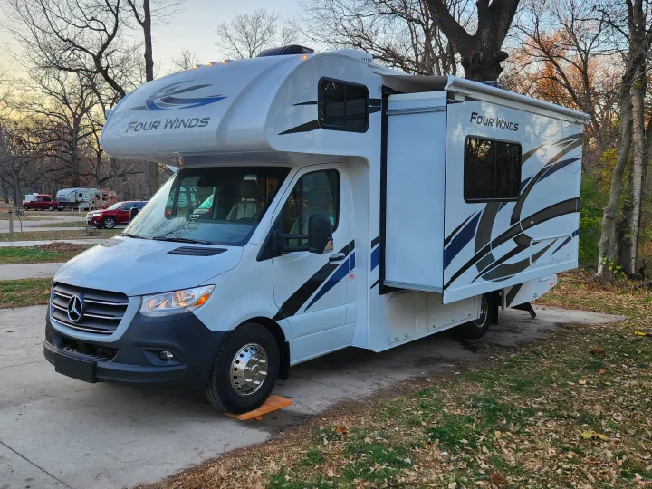 A white Four Winds Class C rv rental, with its slide-out extended, is parked on a concrete pad in a wooded campground.. Thor Motor Coach Four Winds 2023