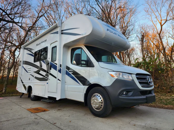 A white Thor Four Winds Class C motorhome, perfect for an rv rental, parked on concrete with trees in the background.. Thor Motor Coach Four Winds 2023