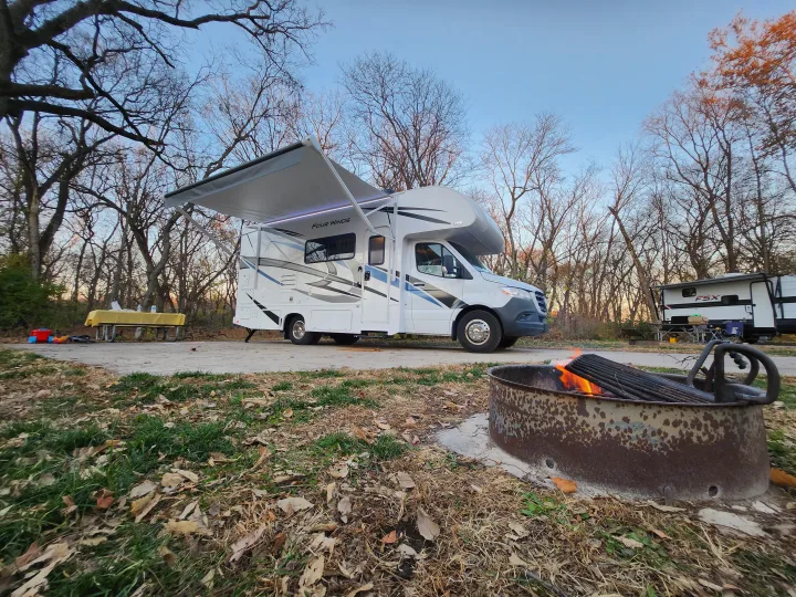 An RV rental, a white Four Winds motorhome, parked at a fall campsite with a glowing fire pit and extended awning.. Thor Motor Coach Four Winds 2023