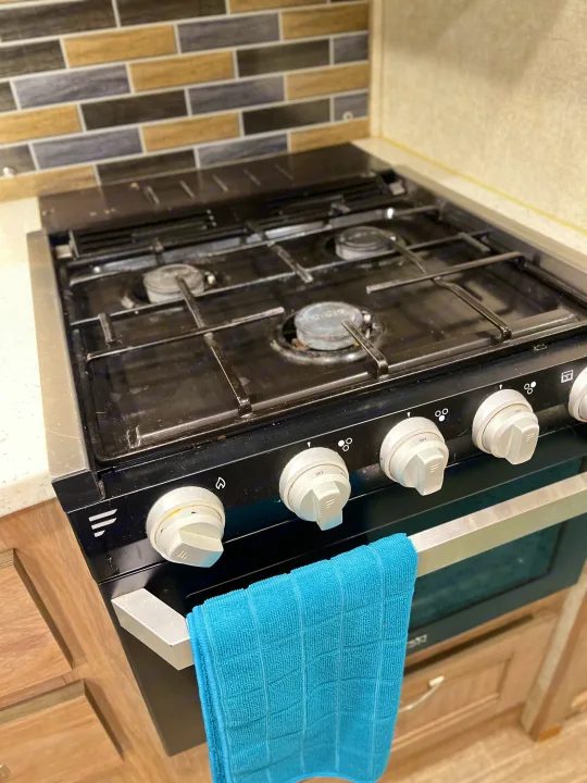 Compact gas stove and oven unit in an RV rental kitchen, featuring a tiled backsplash and blue towel.. Forest River Sunseeker 2019