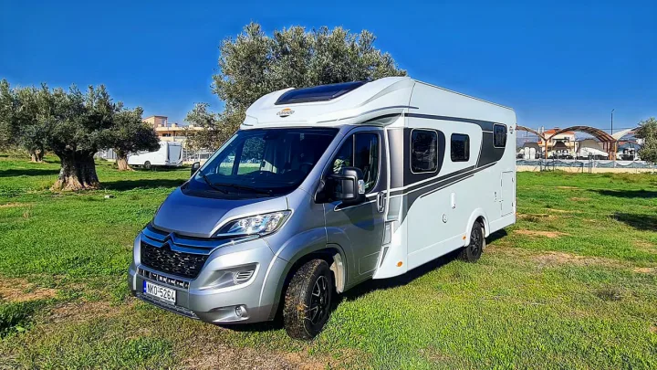 Modern silver & white RV rental on green grass, under clear blue sky, with olive trees in background.. Carado T338 2022