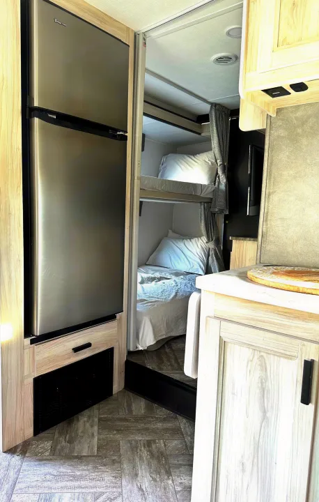 Inside an RV rental: Stainless steel refrigerator and stacked bunk beds with white linens, optimizing space in a cozy mobile home.. Forest River Sunseeker LE 2021