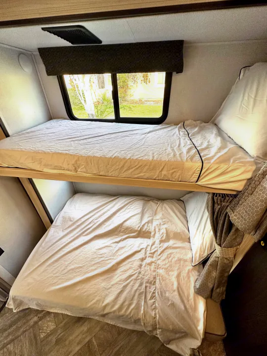 Cozy bunk beds in an RV rental, featuring white sheets, a window view from the top bunk, and a patterned curtain.. Forest River Sunseeker LE 2021