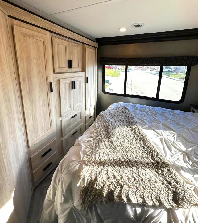 Modern RV rental bedroom: light wood cabinets, large window, comfy bed, and efficient storage. Relax on your trip!. Forest River Sunseeker LE 2021