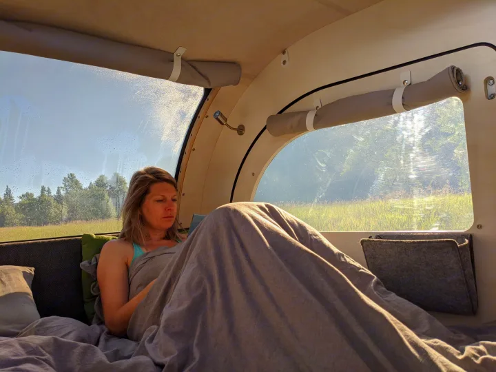 Inside a cozy RV rental: a woman under a blanket, reading with a serene view of trees and fields through the large window.. droplet XL 2026