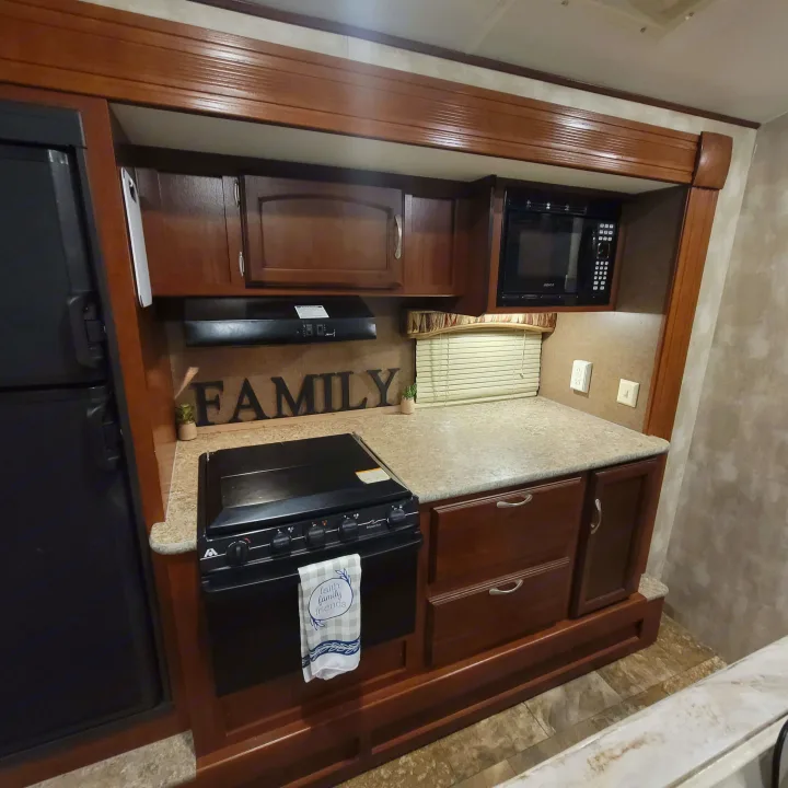 An RV rental's well-equipped kitchen features a black refrigerator, microwave, stove/oven, light countertop, and dark wood cabinets.. Winnebago Ultralite 2014