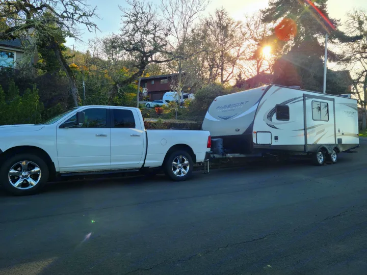 White pickup truck tows a Passport RV, perhaps an RV rental, parked on a sun-drenched street with trees & houses.. Keystone RV Passport Grand Touring 2018