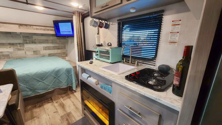 Step into this compact RV rental: featuring a cozy bed, full kitchenette with a turquoise toaster, and an electric fireplace.. Coleman Lantern 2023