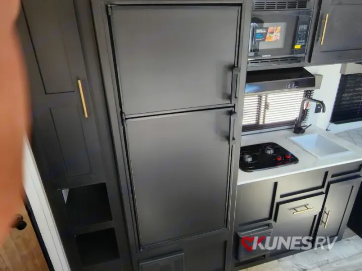 Interior view of a modern RV rental kitchen area, featuring black appliances: a large refrigerator, microwave, sink, and stovetop.. Keystone Springdale 2022