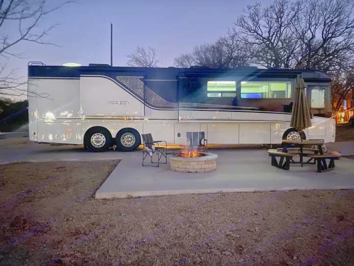 Luxury RV rental at a dusk campsite. Motorhome is parked on a pad with a glowing fire pit, chairs, and picnic table.. Winnebago Horizon 2020