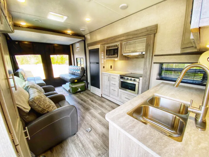 Inside a spacious RV rental: a modern kitchen with double sink, oven, and cozy living area with recliners and wood flooring.. Coachmen Catalina 2022