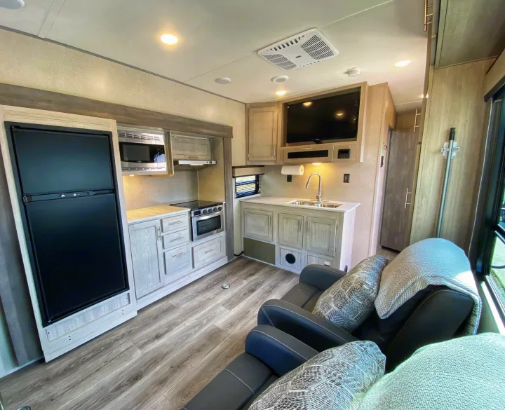 Interior of an RV rental showing a modern kitchen with light cabinets, fridge, stove, sink, and comfy recliners.. Coachmen Catalina 2022