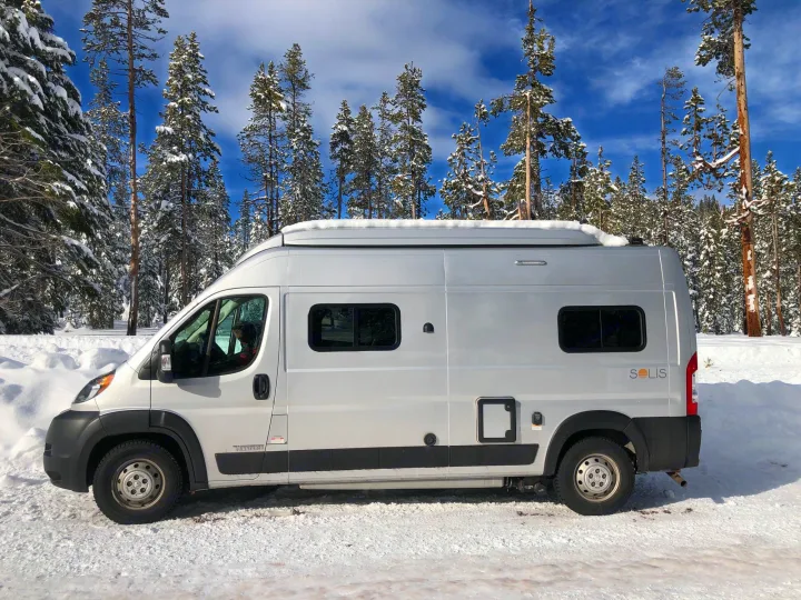 A sleek RV rental, a silver camper van, stands amidst a snowy forest with pine trees under blue skies.. Winnebago Solis 2022