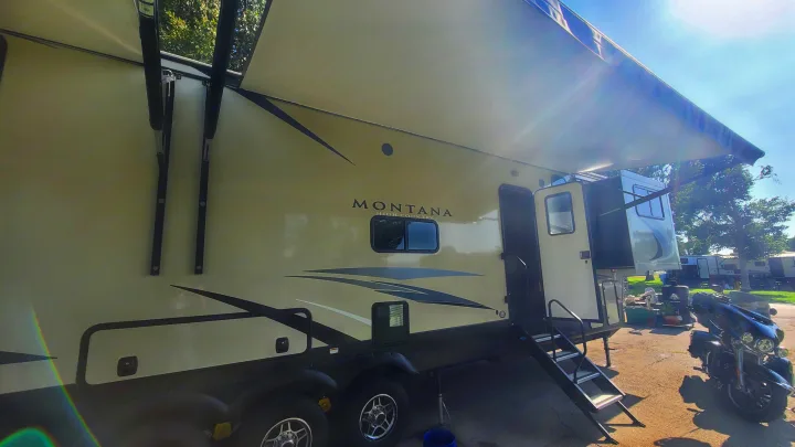 A spacious Montana fifth-wheel RV rental with an extended awning and entry steps, parked beside a motorcycle at a sunny campground.. Keystone RV Montana High Country Toy Hauler 2019