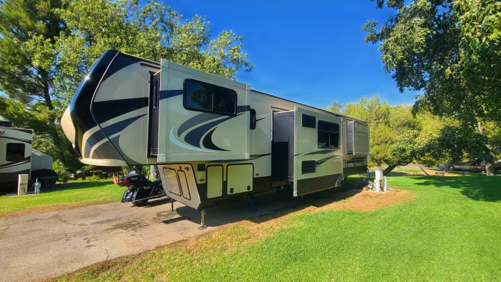 Spacious fifth-wheel RV rental parked with extended slide-outs at a sunny, tree-lined campsite. Perfect for travel!. Keystone RV Montana High Country Toy Hauler 2019